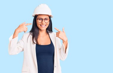 Mimar şapkası takan, gözlüklü neşeli gülümseyen ve parmaklarıyla ve ağzıyla işaret eden genç, güzel bir Latin kız. diş sağlığı kavramı. 