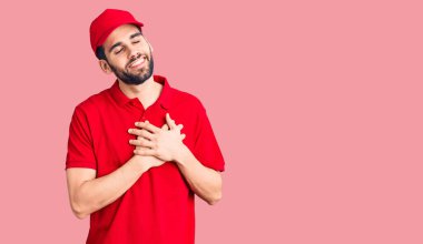 Sakallı, teslimat üniforması giyen, elleri göğsünde, gözleri kapalı ve yüzünde minnet ifadesi olan genç yakışıklı bir adam. sağlık kavramı. 