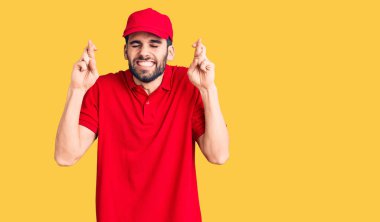 Sakallı, teslimat üniforması giyen, umut dolu ve gözleri kapalı bir şekilde el işareti yapan genç yakışıklı bir adam. Şans ve batıl inançlar. 