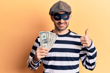 Hırsız maskesi takan genç yakışıklı kel adam dolar çalıyor mutlu ve olumlu gülümsüyor, başparmağıyla mükemmel ve onay işareti yapıyor. 