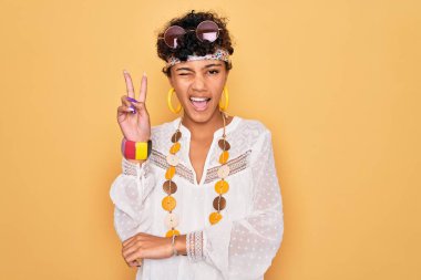 Genç ve güzel Afro-Amerikan Afro-Amerikan kadın güneş gözlüğü takıyor ve aksesuar takıyor. Kameraya gülümsüyor ve parmaklarıyla zafer işareti yapıyor. İki numara..