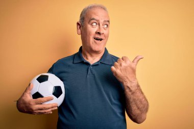 Orta yaşlı beyaz oyuncu futbol topunu sarı zemin üzerinde tutuyor ve gülümseyen bir yüzle baş parmağıyla işaret ediyor.