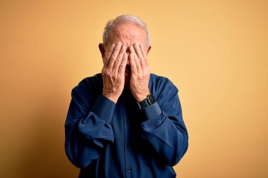 Sarı arka planda dikilen günlük mavi tişört giyen gri saçlı yaşlı bir adam. Ağlarken suratını ellerle kapatan üzgün bir ifade. Depresyon kavramı.