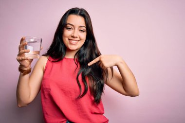 Genç esmer kadın pembe izole edilmiş arka planda bir bardak temiz su içiyor.
