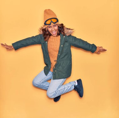 Kışlık giysiler giyen genç, güzel Latin kadın mutlu gülümsüyor. Yüzünde ve kollarında tebessümle, izole edilmiş sarı arka planda atlıyor.