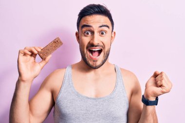 Sakallı, enerji proteini yiyen genç yakışıklı adam pembe arka planda gururla bağırıyor, zaferi kutluyor ve kollarını kaldırarak başarıyı kutluyor.