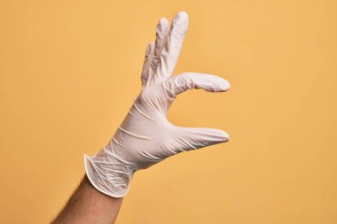 Sarı arka plan üzerinde tıbbi eldiveni olan beyaz bir gencin eli. Görünmez bir şeyi alıp götürüyor. Parmakları yer gösteren bir nesneyi tutuyor.