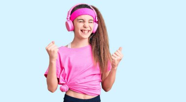 Spor kıyafetleri giyen, kulaklık takan sevimli İspanyol kız çok mutlu ve heyecanlı kollarını kaldırıp, gülümseyerek ve başarı için çığlık atarak kazanan jestler yapıyor. kutlama konsepti. 