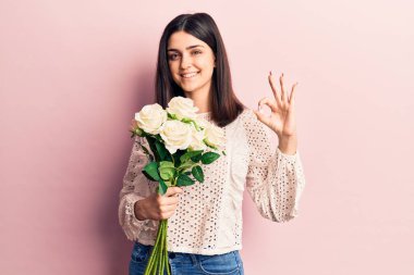 Çiçek buketi tutan güzel bir kız parmaklarıyla imza atıyor, gülümseyerek mükemmel bir sembol oluşturuyor. 