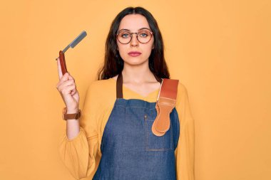 Önlük takan genç ve güzel bir berber kadın sarı arka planda jilet tutuyor. Zeki bir yüz ifadesiyle ciddi ciddi düşünüyor.