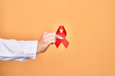 İzole edilmiş sarı arka plan üzerinde HIV hastalığını destekleyen kırmızı kanser kurdelesi tutan beyaz bir kadının eli.