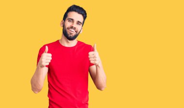 Sakallı, sıradan tişörtler giyen, elinde olumlu jest yapan, gülümseyen ve mutlu genç yakışıklı bir adam. Neşeli ifade ve kazanan jest. 