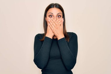 Genç, güzel, esmer, günlük tişört giyen bir kadın izole edilmiş beyaz arka planda dikiliyor. Ağzını yanlışlıkla ellerle kapatıyor. Gizli konsept.