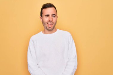 Mavi gözlü, sıradan bir süveter giyen, sarı arka planda duran, kameraya seksi bir ifadeyle bakan, neşeli ve mutlu yüzlü genç bir adam..
