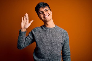 İzole edilmiş turuncu arka plan üzerinde duran genç ve yakışıklı bir adam mutlu ve güler yüzlü, dostça bir jest yapıyor.