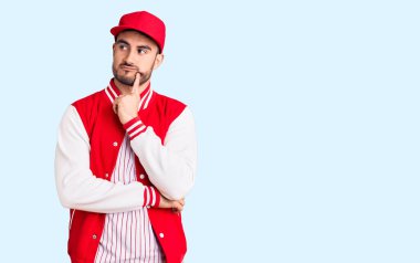 Beyzbol ceketi ve şapka giyen yakışıklı genç adam ciddi bir yüzle eli çenesinde soru sormayı düşünüyor, kafa karıştırıcı bir fikir hakkında düşünceli. 