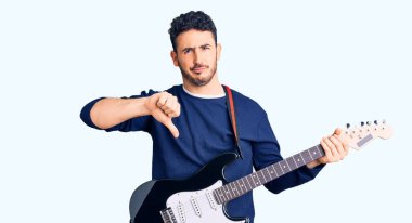 Öfkeli yüzüyle elektro gitar çalan genç İspanyol adam, olumsuz işaret baş parmakları aşağıda, reddedilme konsepti 