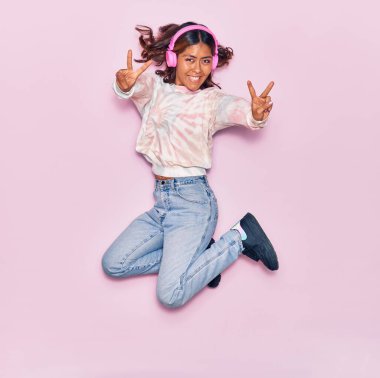 Genç, güzel, Latin bir kadın kulaklık takıp, mutlu bir şekilde müzik dinliyor. Yüzünde gülümsemeyle zıplıyor, pembe arka planda zafer işareti yapıyor.