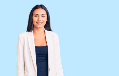 İş elbiseleri giymiş genç, güzel bir Latin kız. Pozitif ve mutlu görünüyor. Dişlerini gösteren bir gülümsemeyle ayakta duruyor ve gülümsüyor. 