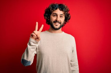 Sakallı, sıradan bir süveter giyen genç, kırmızı arka plan üzerinde duran ve iki numaralı parmaklarıyla işaret eden, kendinden emin ve mutlu gülümseyen bir adam..