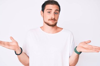 Gündelik beyaz tişört giyen, kolları ve elleri havada, kafası karışmış genç yakışıklı bir adam. şüphe kavramı. 