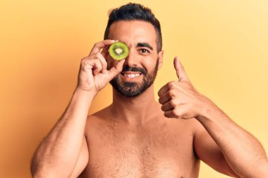 İspanyol asıllı genç adam elinde kivi tutarken mutlu ve pozitif gülümsüyor, başparmağıyla mükemmel ve onay işareti yapıyor. 