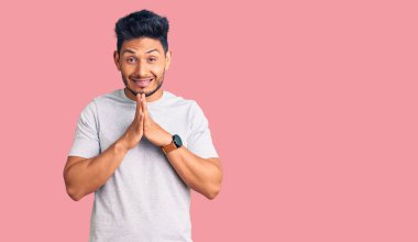 Yakışıklı Latin Amerikalı genç adam gündelik tişört giyip ellerinin birarada dua ederek af diliyor kendinden emin bir şekilde gülümsüyor. 