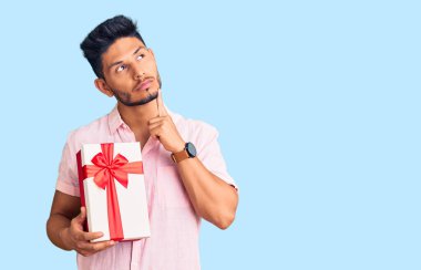 Yakışıklı Latin Amerikalı genç adam elinde hediyeyle ciddi bir yüz ifadesiyle eli çenesinde soru sormayı düşünüyor. Kafa karıştırıcı bir fikir. 