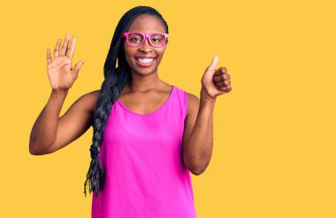 Genç Afro-Amerikalı kadın günlük kıyafetler ve gözlük takıyor. Altıncı parmaklarını gösterip işaret ederken kendinden emin ve mutlu bir şekilde gülümsüyor.. 
