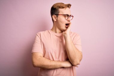 Genç, kızıl saçlı, günlük tişört giyen, izole edilmiş pembe arka planda duran, esnemekten sıkılmış, ağzı eliyle kapatmaktan bıkmış bir adam. Huzursuz ve uykulu..