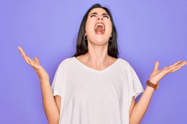 Genç, güzel, esmer bir kadın mor arka plan üzerine gündelik beyaz tişört giyiyor. Deli gibi bağırıyor, agresif bir ifadeyle bağırıyor ve kollarını kaldırıyor. Hayal kırıklığı konsepti.