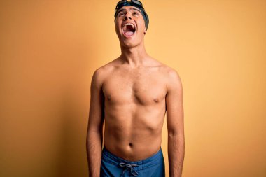 Genç, yakışıklı, mayosuz ve yüzme şapkalı bir adam izole edilmiş sarı arka planda kızgın ve öfkeli bağırıyor, öfkeyle bağırıyor. Öfke ve agresif kavram.