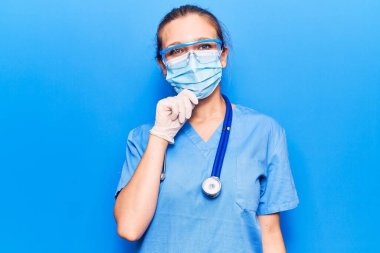 Doktor üniforması ve koronavirüs koruması giyen genç sarışın kadın kameraya çapraz kollar ve çene üzerinde kendine güvenen gülümsemeyi hatırlatır. Olumlu düşünmek. 
