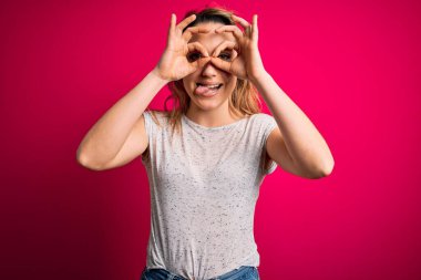 İzole edilmiş pembe arka plan üzerinde sıradan tişörtler giyen genç, güzel sarışın bir kadın dürbünün dili dışarı çıkarması, gözlerin parmaklarından içeri bakması gibi güzel hareketler yapıyor. Çılgın ifade.