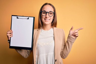 Sarı arka planda kontrol panosu tutan gözlüklü güzel bir müfettiş kadın. Elini ve parmağını kenarı göstererek çok mutlu.