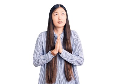 Genç, güzel, Çinli bir kadın gündelik tişörtler giyip elleri bağlı yalvarıyor ve dua ediyor. Yüzünde umut ifadesi var. Çok duygusal ve endişeli. Yalvarıyor. 