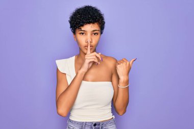 Güzel, Afro-Amerikan Afro-Amerikan bir kadın mor arka plan üzerine gündelik tişört giyip dudaklarını yan tarafı gösteren parmaklarıyla sessiz olmak istiyor. Sessizlik ve gizli konsept.