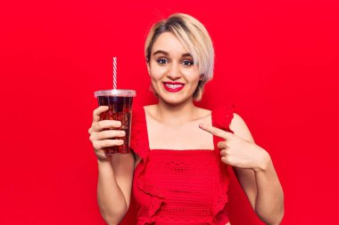 Genç, güzel, sarışın kadın bir bardak kola içiyor gülümsüyor, mutlu bir şekilde elini ve parmağını gösteriyor. 