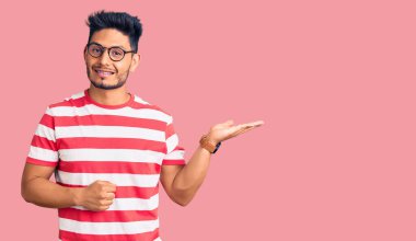 Yakışıklı Latin Amerikalı gündelik kıyafetler ve gözlük takan neşeli bir şekilde gülümseyen ve avuç içi kameraya bakan genç adam.. 