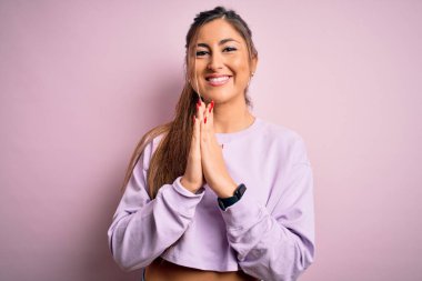 Pembe izole edilmiş arka plan üzerine kazak giyen genç ve güzel bir kadın ellerinin birarada dua ederek af diliyor kendinden emin bir şekilde gülümsüyor..