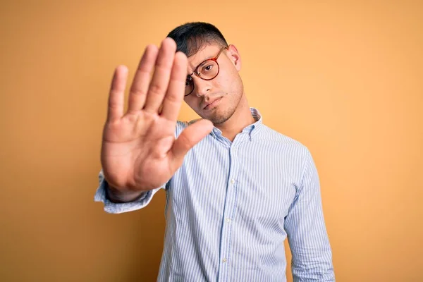 黄色の背景にオタク眼鏡をかけた若いハンサムなヒスパニック系のビジネスマンが手の手のひらで歌うのをやめる 顔に負と深刻なジェスチャーで警告式 — ストック写真