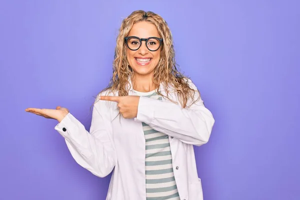 Young beautiful blonde scientist woman wearing coat and glasses over purple background amazed and smiling to the camera while presenting with hand and pointing with finger.
