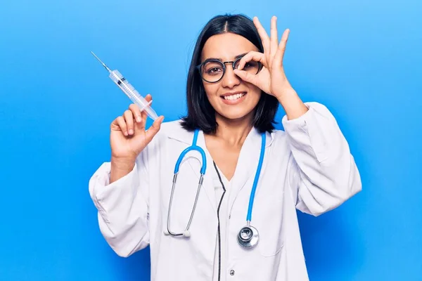 Young beautiful latin woman wearing doctor stethoscope holding syringe smiling happy doing ok sign with hand on eye looking through fingers