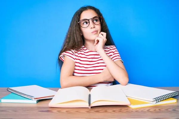 Cute hispanic child girl studying for school exam sitting on the table thinking concentrated about doubt with finger on chin and looking up wondering