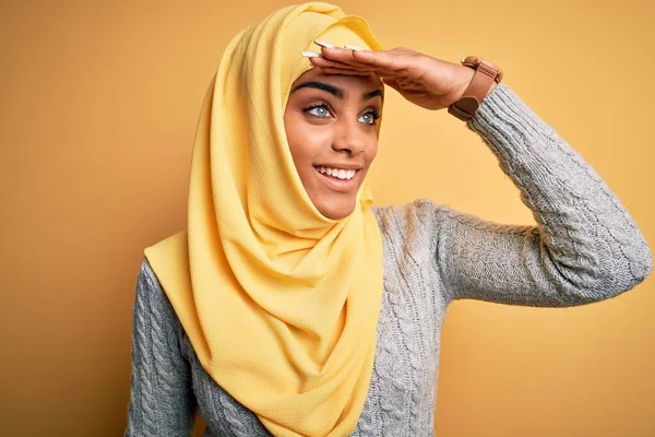 Young beautiful african american girl wearing muslim hijab over isolated yellow background very happy and smiling looking far away with hand over head. Searching concept.