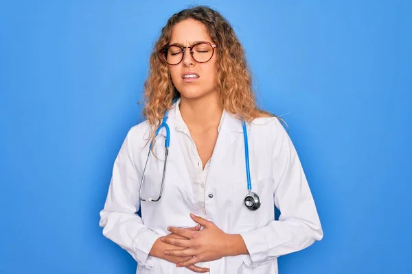 Young beautiful blonde doctor woman with blue eyes wearing coat and stethoscope with hand on stomach because nausea, painful disease feeling unwell. Ache concept.