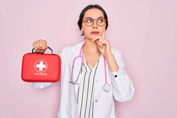 Young beautiful doctor woman with blue eyes wearing stethoscope holding first aid kit box serious face thinking about question, very confused idea
