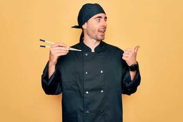 Young handsome cooker man with blue eyes wearing uniform and hat using chopsticks pointing and showing with thumb up to the side with happy face smiling