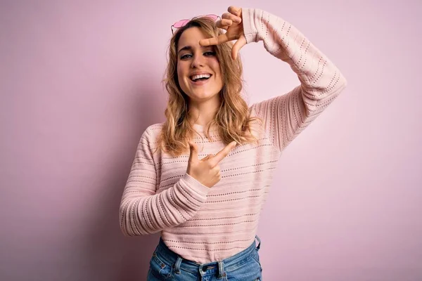ピンクの背景にカジュアルなセーターとサングラスを身に着けている若い美しいブロンドの女性は 幸せな顔で手や指でフレームを作り笑顔 創造性と写真の概念 — ストック写真