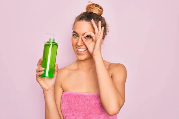 Beautiful blonde woman with blue eyes wearing towel shower after bath holding aloe vera gel with happy face smiling doing ok sign with hand on eye looking through fingers